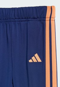 Námornícke modré športové nohavice s elastickým pásom, bočnými vreckami a oranžovým logom Adidas a pruhmi po stranách.