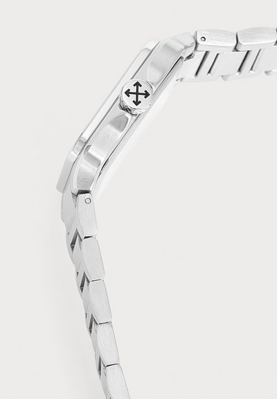 Vue latérale d'une montre-bracelet en métal argenté avec un bracelet maillé et une couronne ornée d'un symbole de flèche croisée.