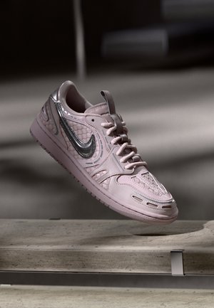 Jasnoróżowy sneaker Nike z srebrnym logo swoosh i teksturowanymi detalami, unoszący się nad betonową krawędzią na ciemnym, rozmytym tle.