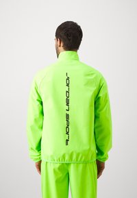 Neon zelena lahka jakna z visokim ovratnikom in elastičnimi manžetami, ki ima na hrbtni strani vertikalni črni logo "Jordan Sport".