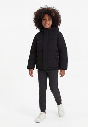Enfant aux cheveux bouclés souriant et marchant, portant une doudoune noire, un pantalon foncé et des baskets blanches sur fond blanc.
