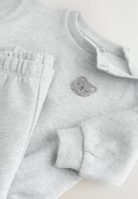 Ljust grå babyoutfit med långa ärmar, byxor med elastisk midja, tryckknappar på axeln och en liten texturerad björnpatch på bröstet.