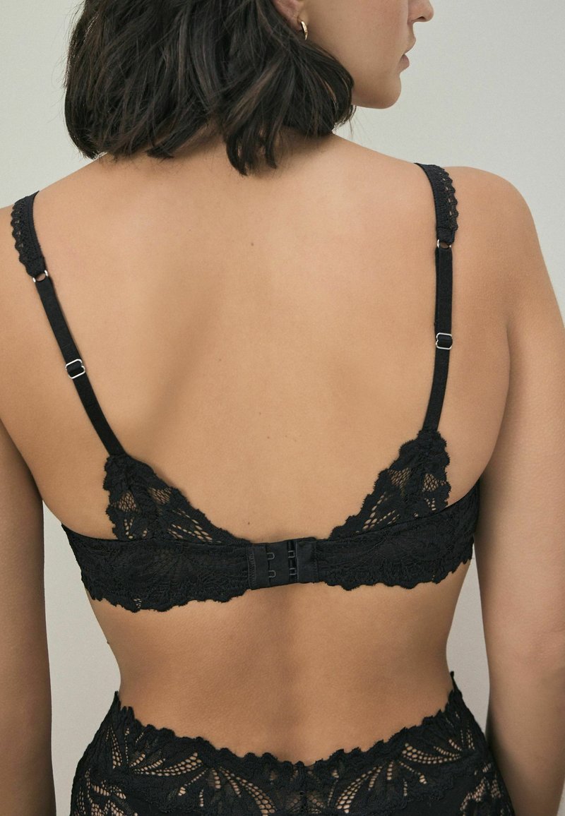 Bralette de encaje negro con bordes en scallop, tirantes ajustables y cierre posterior de gancho y ojo con patrón floral decorativo.