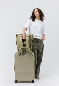 Uno zaino verde oliva con spallacci regolabili poggia su una valigia beige con ruote. Entrambi gli articoli presentano un design liscio e robusto con hardware funzionale.