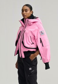 Nő rózsaszín kapucnis dzsekiben, fekete cipzárokkal és fekete nadrágban, oldalzsebekkel, egyik kezét a zsebében tartva, egyszínű háttér előtt áll.