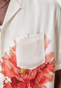 Camisa de manga corta con estampado floral en naranja y crema, con un bolsillo en el lado izquierdo y botones beige en la parte delantera.