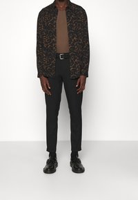 Musta paita, jossa ruskea abstrakti kuvio, ruskea aluspaita, mustat housut ja mustat kengät. Kapea malli, jossa napitettu kaulus ja vyö.