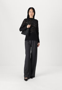 Black lace long-sleeve top with floral pattern, paired with gray wide-leg trousers and black handbag. Model stands against a white background.