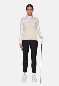 Hellbeige Reißverschluss-Jacke mit weißen Streifen, kombiniert mit schwarzen, enganliegenden Hosen. Accessoires umfassen einen Golfschläger und einen Handschuh, ergänzt durch weiße Sneaker.