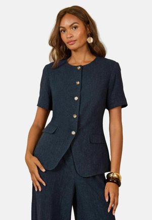 Woman wearing a dark blue short-sleeve buttoned blazer and matching wide-leg pants with gold and brown bangles on her wrist.