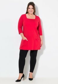 Rote Tunika mit quadratischem Ausschnitt, Dreiviertelärmeln und Fronttaschen, kombiniert mit schwarzen Leggings und schwarzen Absätzen. Glatte Textur.