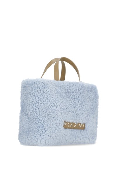 Borsa in pelliccia sintetica azzurro chiaro con un esterno morbido e testurizzato. Presenta manici beige e un'etichetta con logo dorato. Forma rettangolare con bordi arrotondati.
