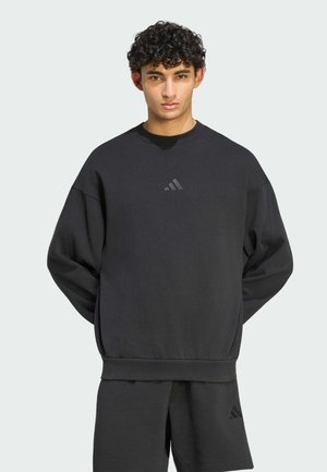 Jeune homme aux cheveux bouclés portant un sweat noir Adidas et un short assorti, debout devant un fond uni clair.