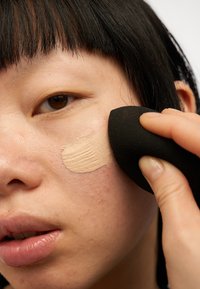Makeup svamp påfører foundation på huden. Svampen er sort; foundation-fargen er beige. Jevn hudtekstur med lette ujevnheter synlige.