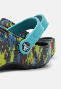 Des sabots colorés avec une base noire, éclaboussures de bleu, vert et jaune, dotés d'une sangle ajustable en sarcelle et d'un détail de logo rond.