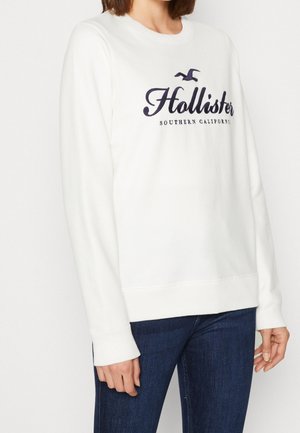 Persoon draagt een witte Hollister-trui met lange mouwen met marineblauwe tekst en logo, gecombineerd met donkerblauwe jeans tegen een effen achtergrond.