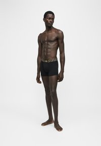 Homme grand à la peau foncée, debout pieds nus, portant un boxer noir avec une ceinture dorée à motifs, sur un fond blanc uni.