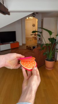 Oranžinės ir raudonos plastikinės konteinerio formos, primenančios vaisius su baltomis sėklomis, sukrautos ir laikomos rankose; lygi tekstūra, išlenktas dizainas, žaisminga išvaizda.