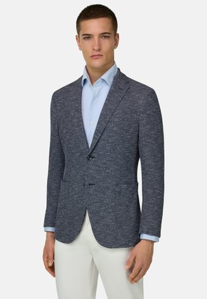 Jonge man staand, gekleed in een blauw visgraatblazer over een lichtblauw overhemd met witte broek, tegen een effen lichte achtergrond.
