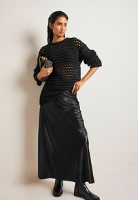 Pulover negru din tricot deschis, asortat cu o fustă maxi din satin negru și cizme cu toc negre; ținuta are un model texturat și o geantă tip clutch.