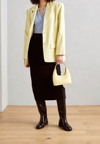 Gelber oversized Blazer über einem hellblauen Oberteil, schwarzer Bleistiftrock und knöchelhohe braune Stiefel. Hält eine kleine gelbe Handtasche.