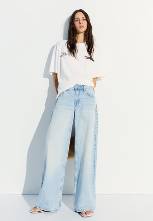 Tânără cu păr lung și închis la culoare, purtând un tricou alb larg și blugi albaștri deschiși, largi, stând în picioare în fața unui perete alb.