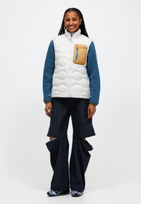 Fuzzy vit väst med en tanfärgad dragkedjeficka, blå strukturerade ärmar och breda svarta byxor med sprickor. Utomhusskor med ljusblå detaljer.