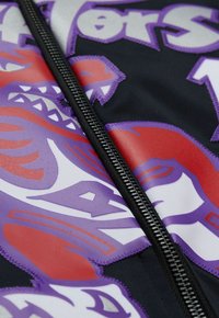 Giacca nera con zip caratterizzata da audaci grafiche rosse e viola. Texture liscia con cuciture in evidenza lungo la zip. I motivi includono un motivo animale feroce.