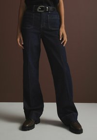 Jeans ampi in denim scuro con vita alta, cuciture a contrasto e tasche anteriori e posteriori, abbinati a una cintura nera e scarpe marroni.