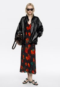 Frau trägt schwarze Lederjacke über langem Blumenkleid, schwarze Sonnenbrille, hält dunkelbraune Tasche, steht in mehrfarbigen offenen Sandalen vor weißem Hintergrund.