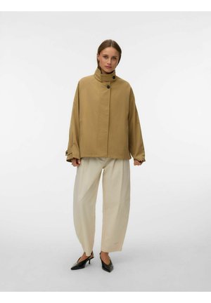 Kvinde iført en løs tanjakke med høj krave, brede beige bukser og sorte spidse hæle, stående mod en ensfarvet hvid baggrund.