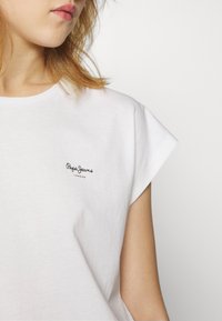 Fehér, rövid ujjú pamut póló kerek nyakkal. A bal mellen egy kis fekete "Pepe Jeans London" logó található.