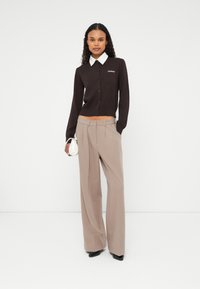 Cardigan marron avec col blanc, fermeture à boutons et logo. Associé à un pantalon beige à jambes larges et des chaussures à talons noires. Tenant un petit sac blanc.