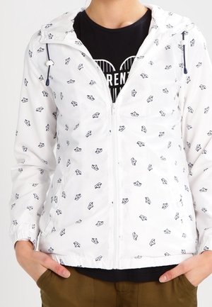 Witte jas met ritssluiting met zwarte papieren bootpatronen, gedragen over een zwart shirt, gecombineerd met een bruine broek, handen in de zakken.