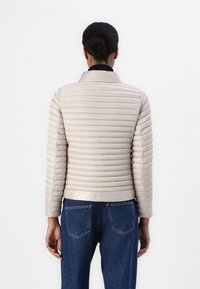 Lyse beige pufferjakke med vandret syninger, stående krave og ribbede manchetter. Parret med mørkeblå jeans, set fra ryggen.