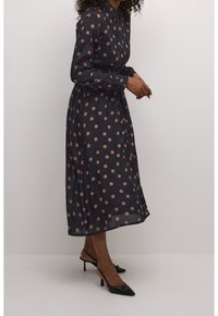 Marinblå, långärmad klänning med stora beige prickar, knappstängning framtill, bälte i midjan och en flödande silhuett. Svarta spetiga klackskor.