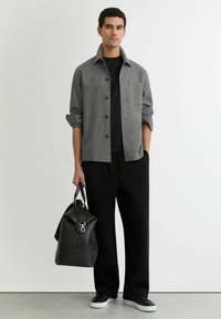 Grijze overhemd met knopen en een kraag, zwarte T-shirt met ronde hals eronder, zwarte broek en een zwarte leren tas met een gouden accent. Witte sneakers.