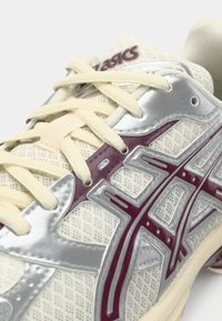 Μπεζ αθλητικό παπούτσι με πλέγμα στην επάνω πλευρά, ασημένιες και πορφυρές λεπτομέρειες και υφή. Διαθέτει κρέμα κορδόνια και ενισχυμένο καπάκι δαχτύλων.