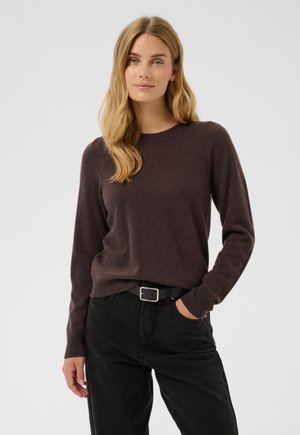 Jeune femme aux longs cheveux châtain clair portant un pull marron foncé et un pantalon noir avec une ceinture noire, posant avec une main dans la poche.