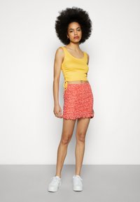Top cropped amarelo com textura em canelado e franzido lateral, combinado com uma saia mini de estampa floral vermelha com bainha volumosa. Usado com ténis brancos.