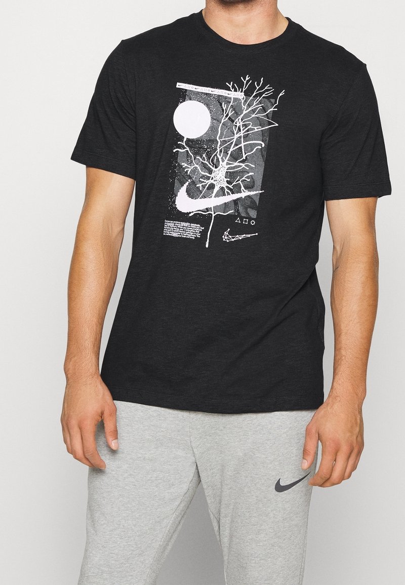Man som bär en svart Nike-t-shirt med vitt abstrakt neuron-mönster och vit Nike-logotyp, tillsammans med ljusgrå mjukisbyxor med svart Nike-logotyp.