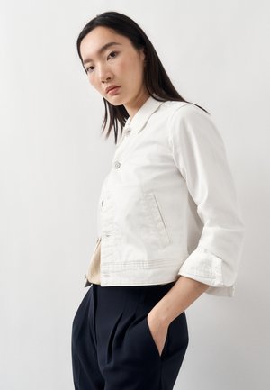 Žena s dlhými tmavými vlasmi, oblečená v bielom zapínanom saku a tmavých plisovaných nohaviciach, stojaca s jednou rukou v vrecku na bielom pozadí.