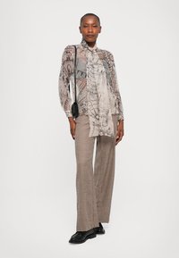 Bluză transparentă bej cu imprimeu arhitectural și detalii cu fundiță, purtată împreună cu pantaloni largi cu model houndstooth în nuanțe pământii. Pantofi și geantă negre.