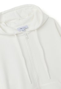 Sudadera blanca con cremallera hecha de una tela suave. Presenta una capucha con cordones, bolsillos frontales y una textura lisa con detalles de costura sutiles.
