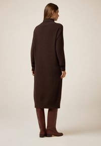 Vestido de punto marrón con un cuello alto, mangas largas y puños acanalados. El vestido llega por debajo de la rodilla, combinado con botas marrones hasta la rodilla.