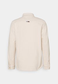 Camicia beige a maniche lunghe vista da dietro con un piccolo logo rosso, bianco e blu vicino al colletto e polsini abbottonati.