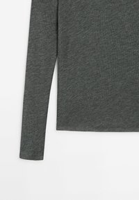 Manche longue de chemise gris foncé et ourlet latéral inférieur sur fond blanc, montrant la texture du tissu et les détails de couture.