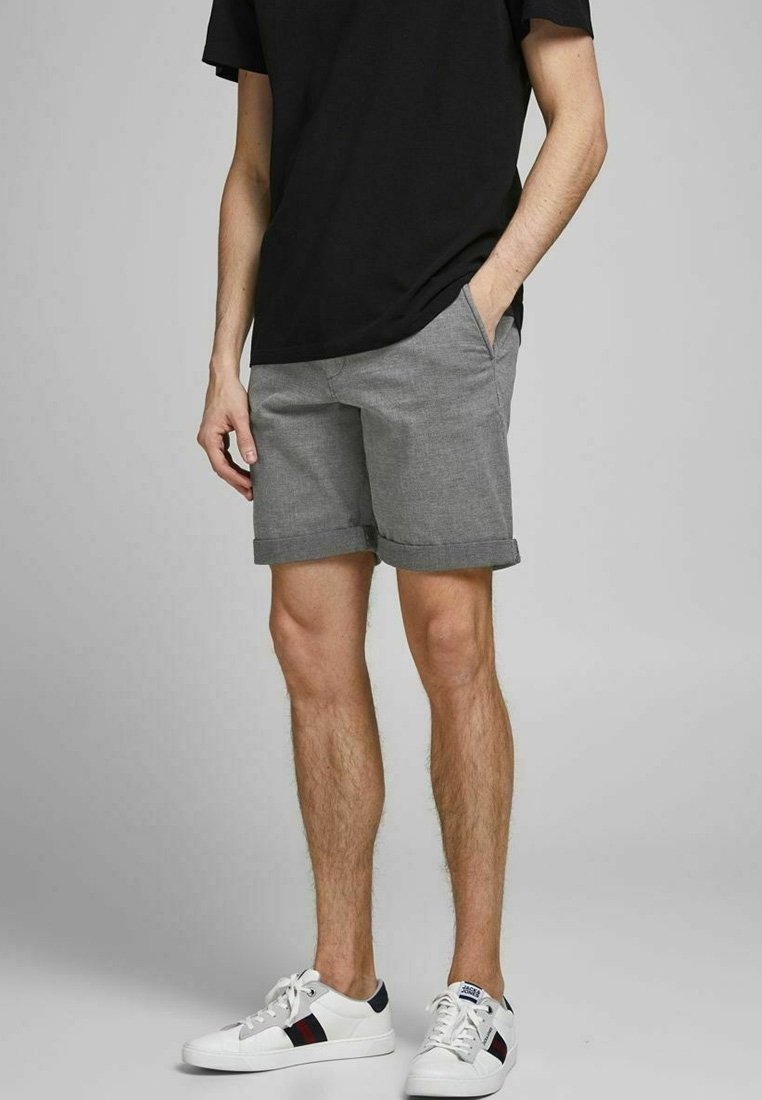Shorts gris avec revers roulés, en tissu texturé. Associés à une chemise noire et des baskets blanches avec un accent de couleur.