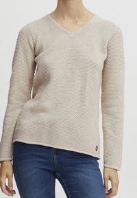 Pull beige clair texturé avec un col en V et des manches longues, présentant un motif de tissage distinctif et une petite étiquette de marque à l'ourlet.