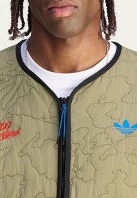 Oliwkowa pikowana kurtka bomberka z czarnymi wykończeniami, z niebieskimi akcentami zamka błyskawicznego i niebieskim haftowanym logo oraz czerwonym haftowanym tekstem.